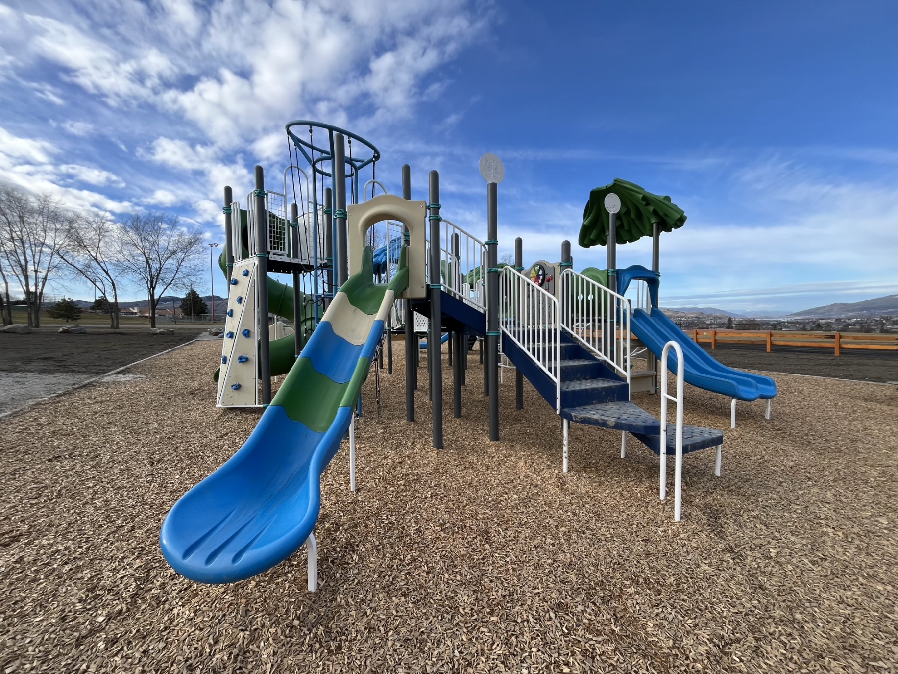 Striped slide, stairs and double slide at DND Grounds Playground in Vernon, BC, beside a wooden fence.
