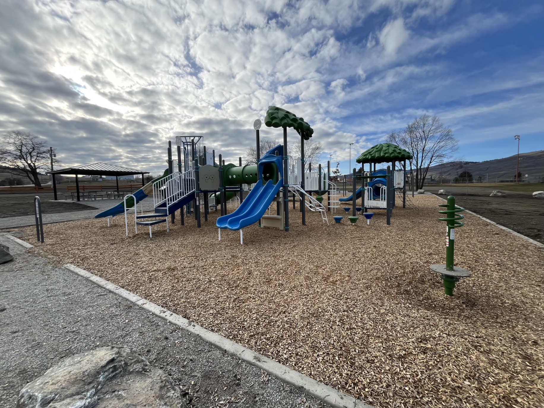 DND Grounds Playground in Vernon, BC, with tree canopy towers, blue slides and a picnic shelter.