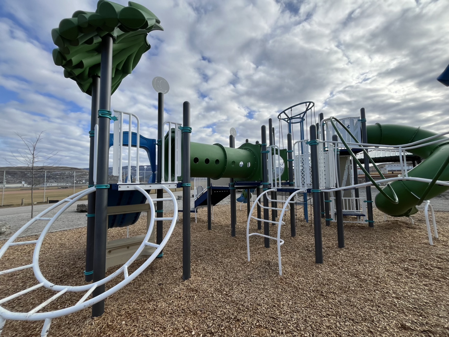 White loop ladder and green crawl tunnel on the play structure, with hills in the distance.