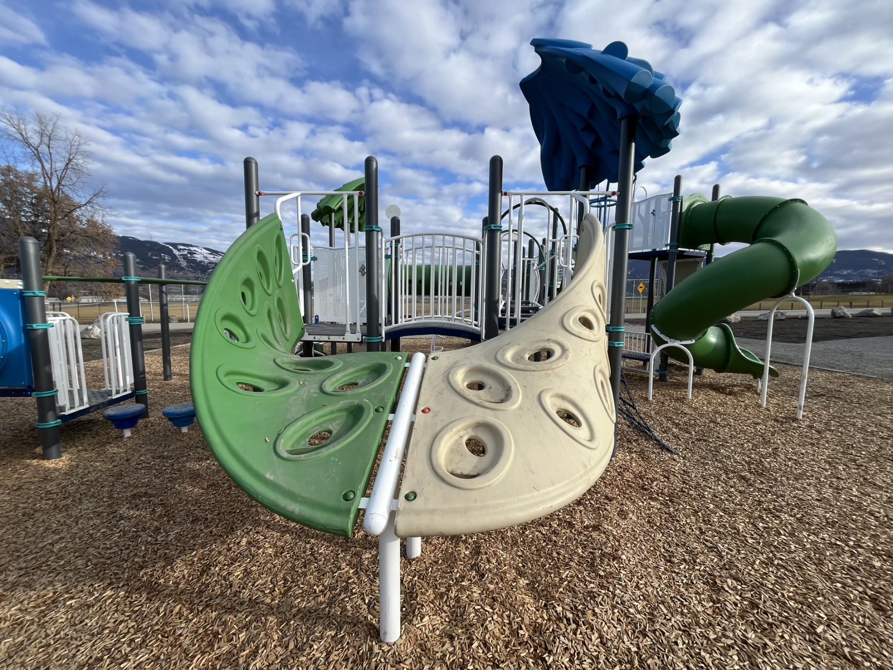Curved green and beige climbing wall at DND Grounds Playground in Vernon, BC, snowy mountains behind.
