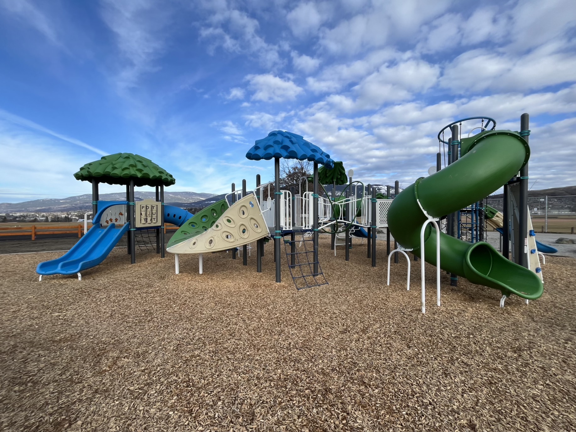 Wide view of DND Grounds Playground in Vernon, BC, with a climbing wall and green spiral slide.