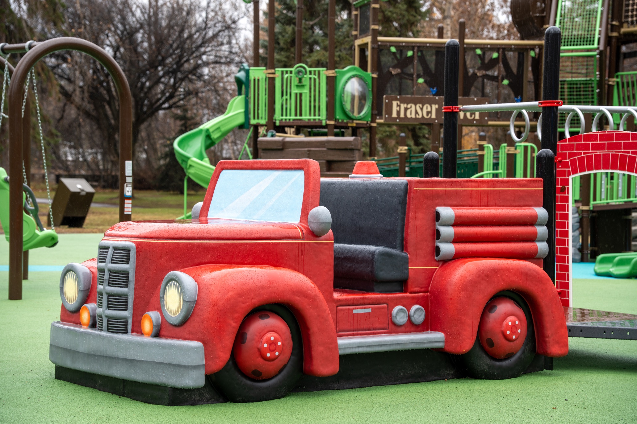Red fire truck sculpture on green rubber surfacing, with a green spiral slide and swings behind.