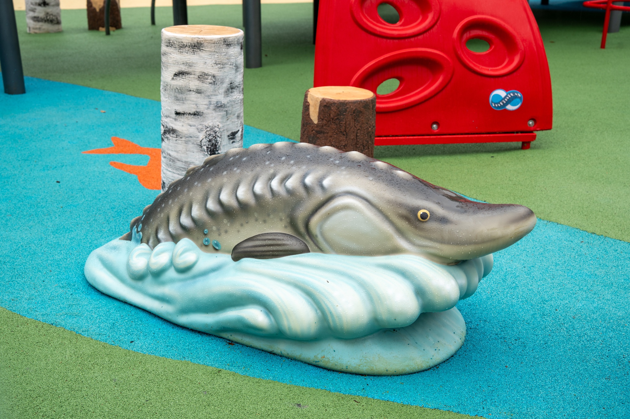 Sturgeon sculpture on a wave base, surrounded by birch log posts and a red climbing wall.