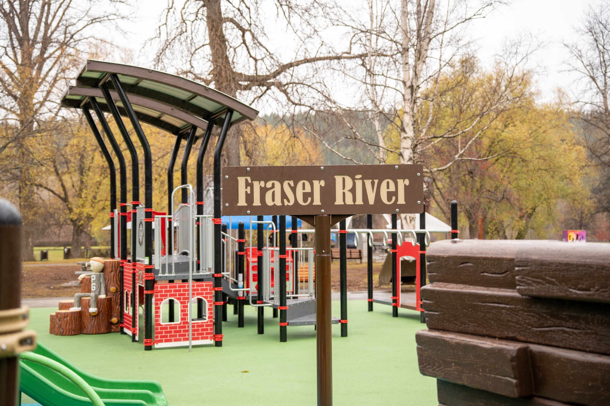 Fraser River sign in front of a red brick-panel play structure with curved canopies and autumn trees.