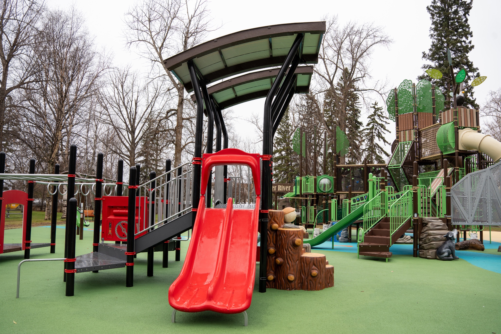 Red double slide under a curved canopy, with the tree-themed tower and a wolf sculpture behind.
