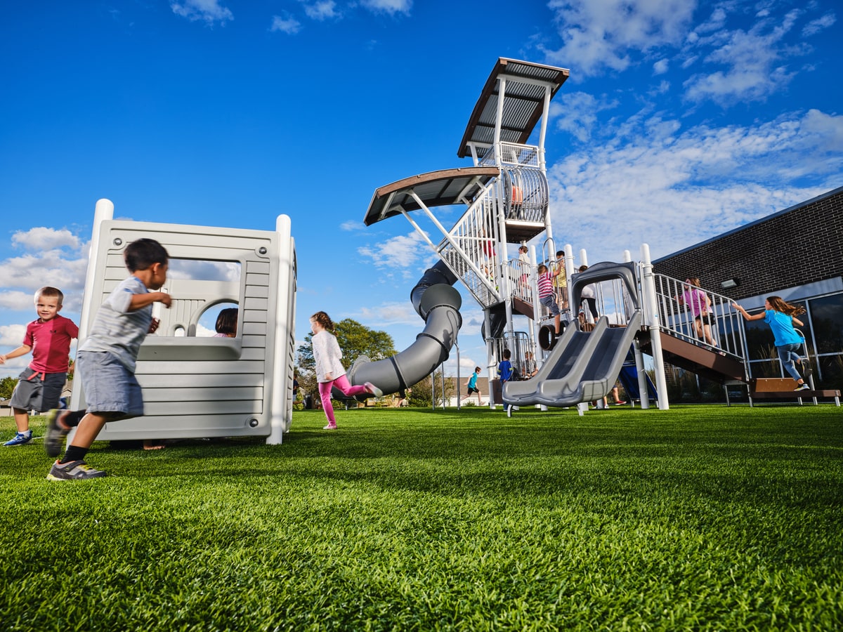 Children running across grass toward a grey play tower with a tube slide and stairs.