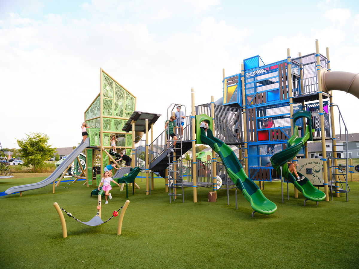 Kids climbing and sliding on a large green and tan commercial playground design set on artificial turf.
