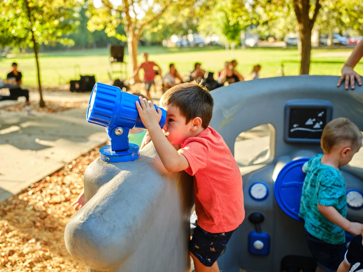 Child looking through a telescope panel on a play structure, another child beside a steering panel.