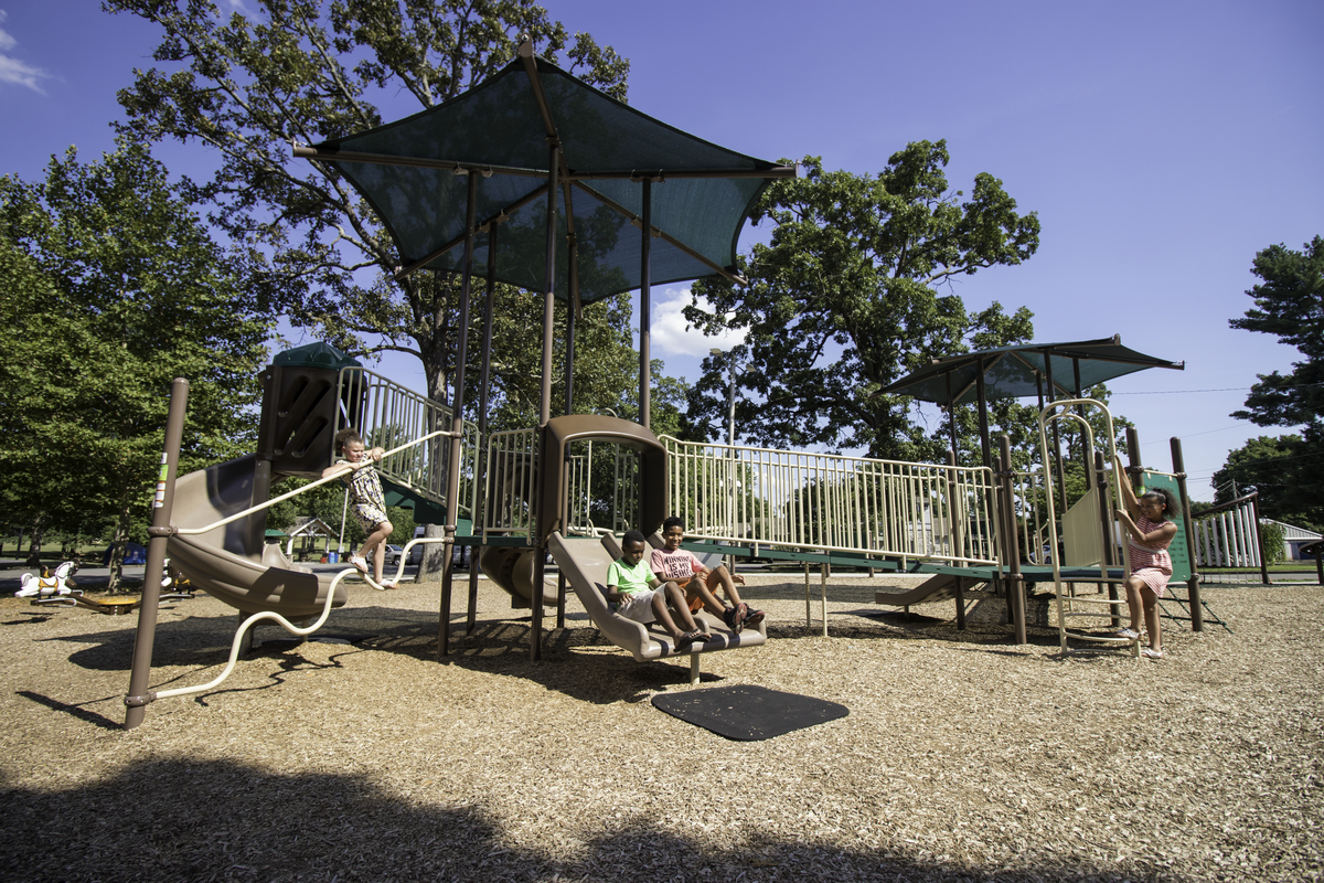 Children sliding and climbing on a brown play structure shaded by canopies over a wood chip safety surface.
