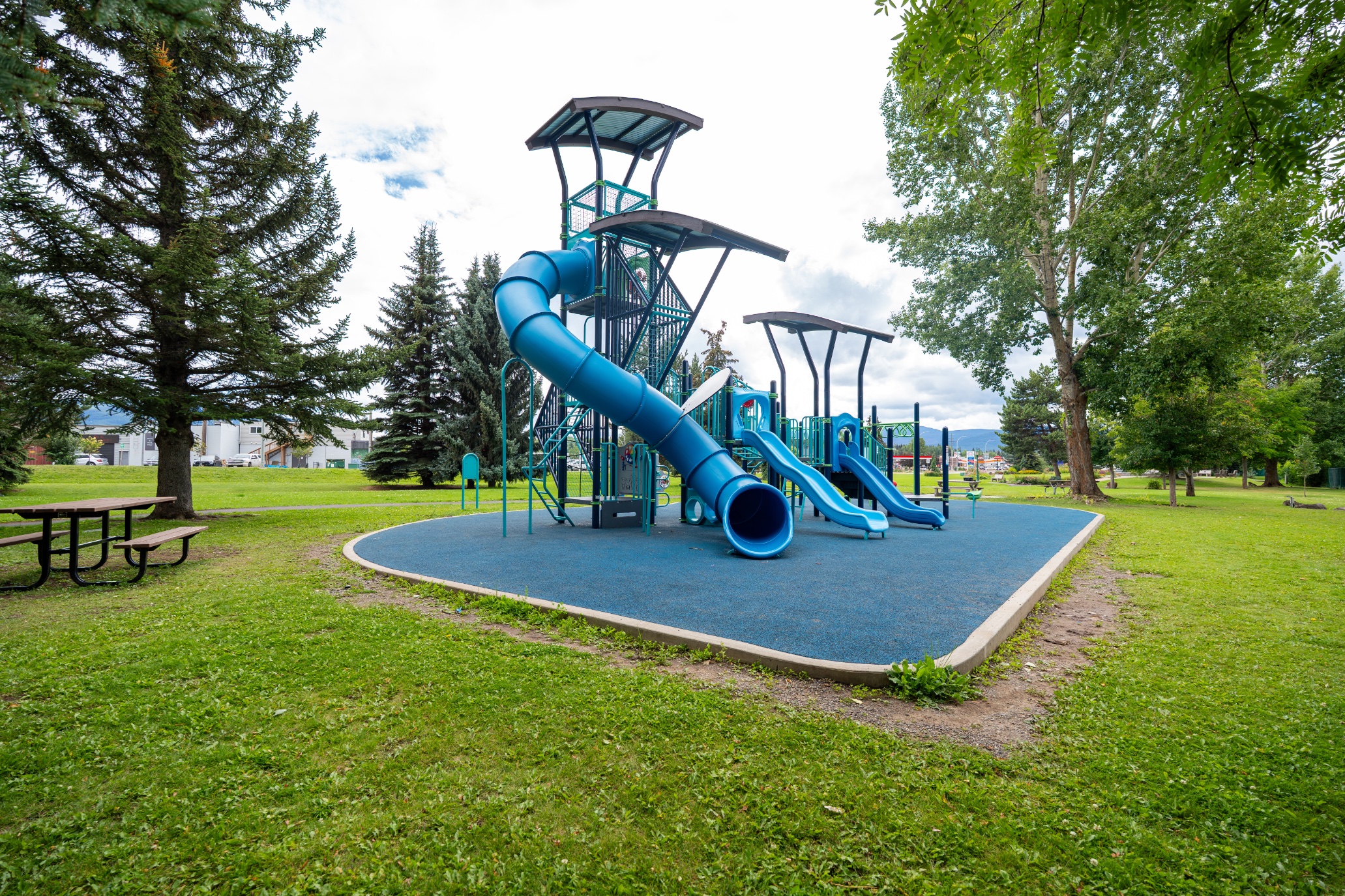 Tall tube slide and shaded play tower on blue rubber surfacing beside a park picnic table.