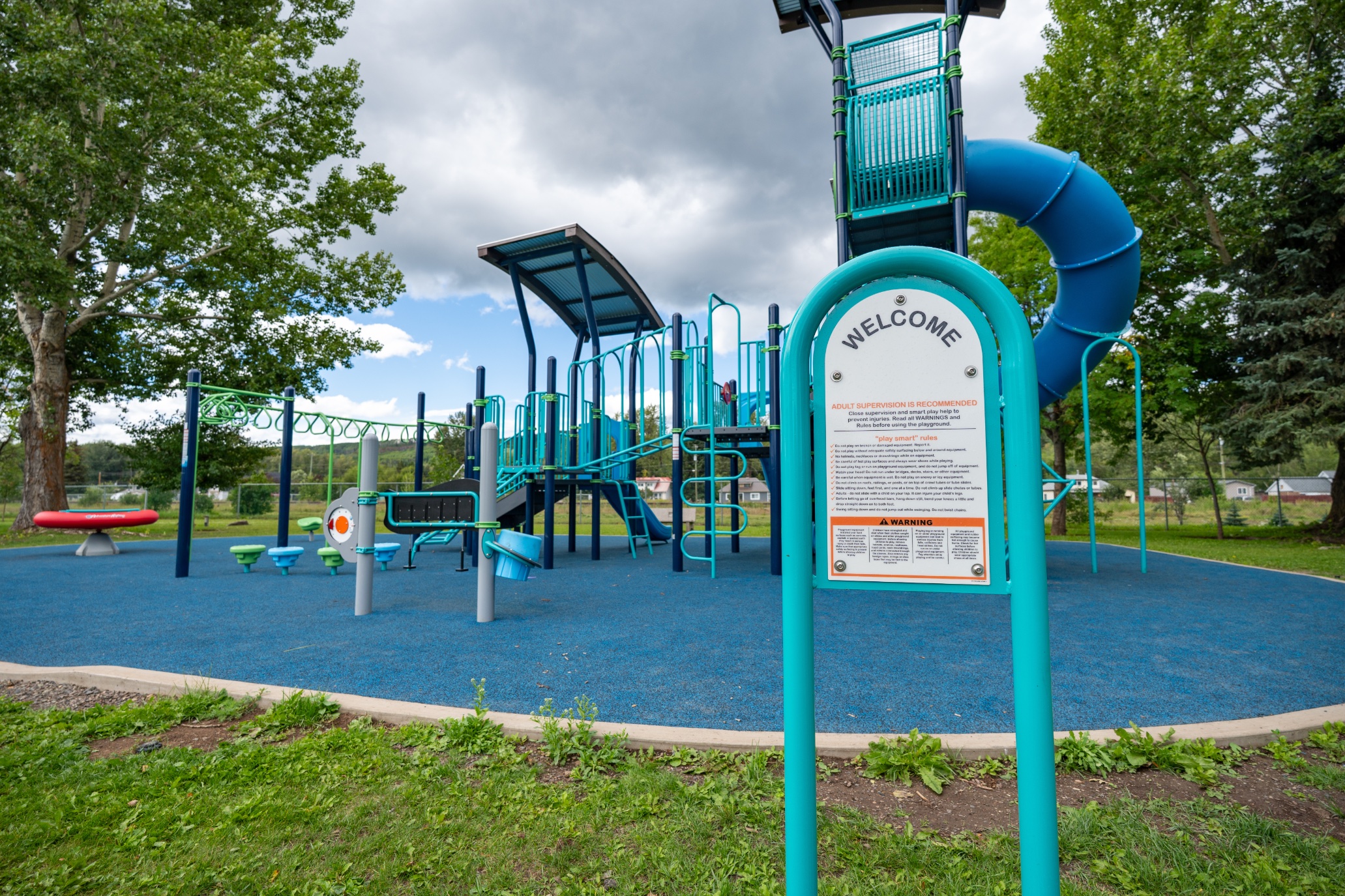 Welcome and play-safe rules sign at the entrance to a blue rubber-surfaced playground.