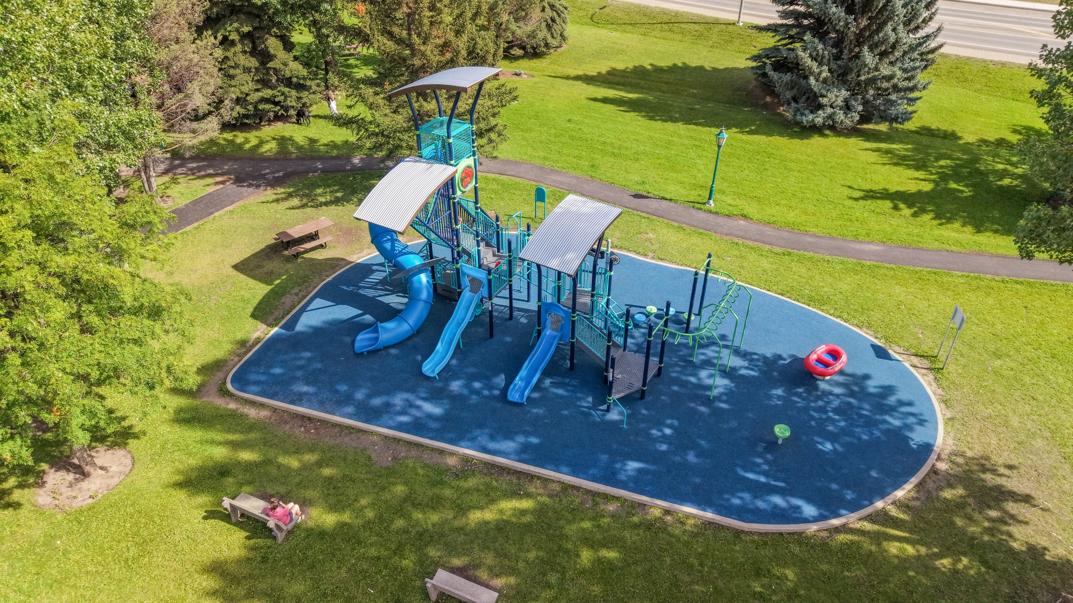 Overhead view of an oval blue play area with three slides, shade canopies and a paved walking path.