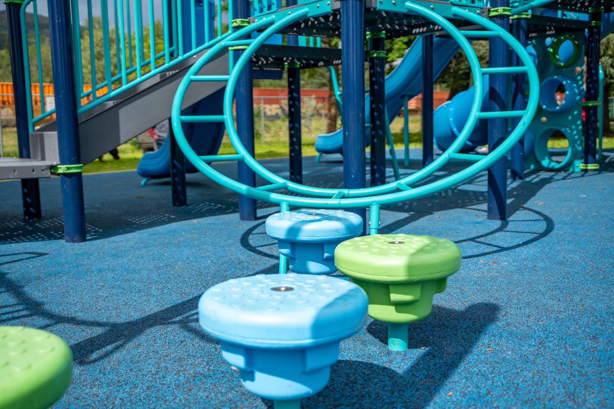 Teal circular ring climber and three stationary stepping seats on blue rubber safety surfacing.