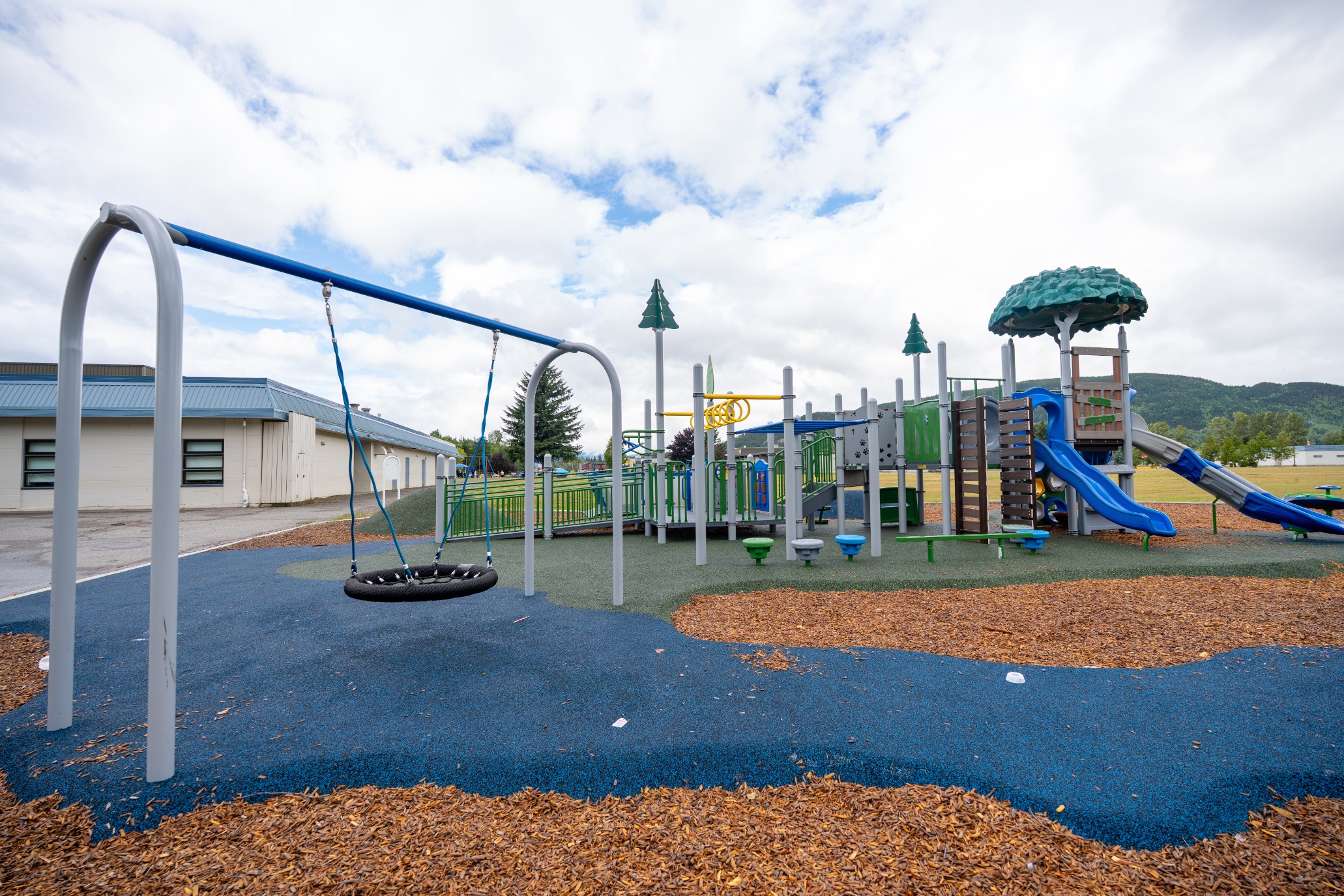 Playground with a basket swing, green tower, blue slides and monkey bars, mountains in the background.