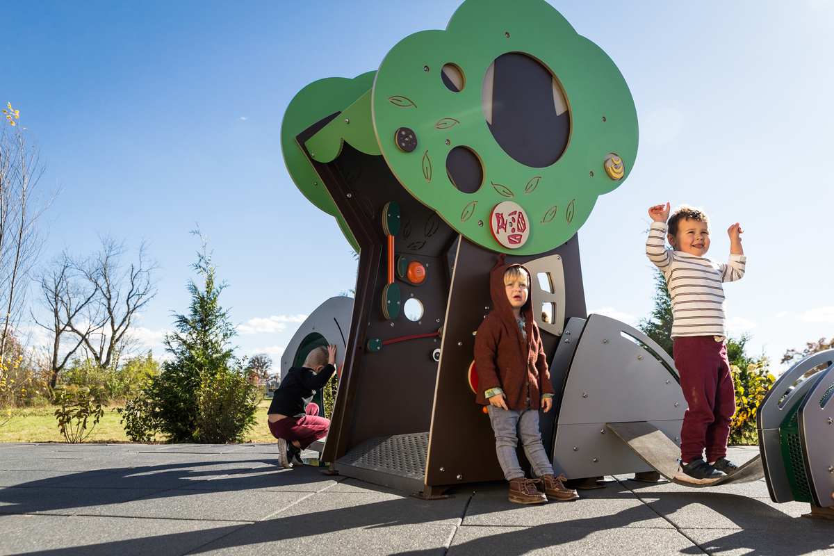 Three toddlers playing at a tree-shaped activity panel structure on rubber tile surfacing.