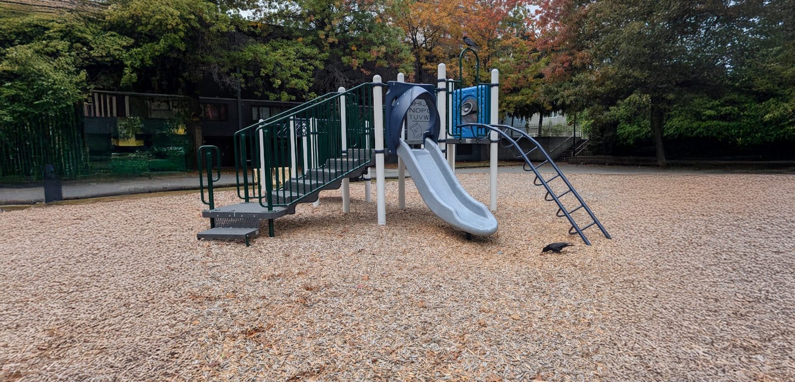 Grey slide, curved ladder and alphabet panel on the wood chip playground at MacLean Park in Vancouver, BC.