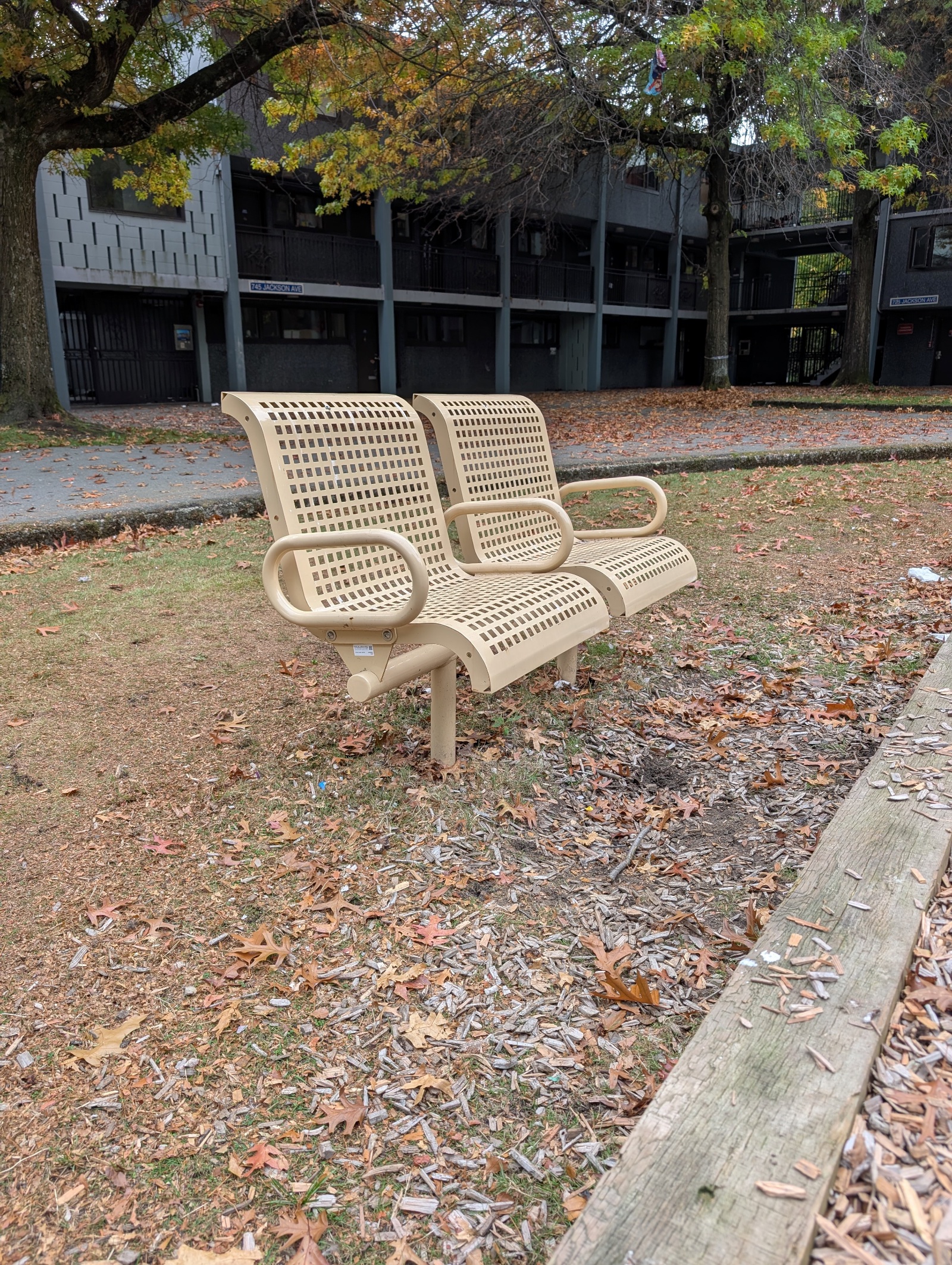 Tan perforated double bench seating near the playground, with a low-rise apartment building behind.