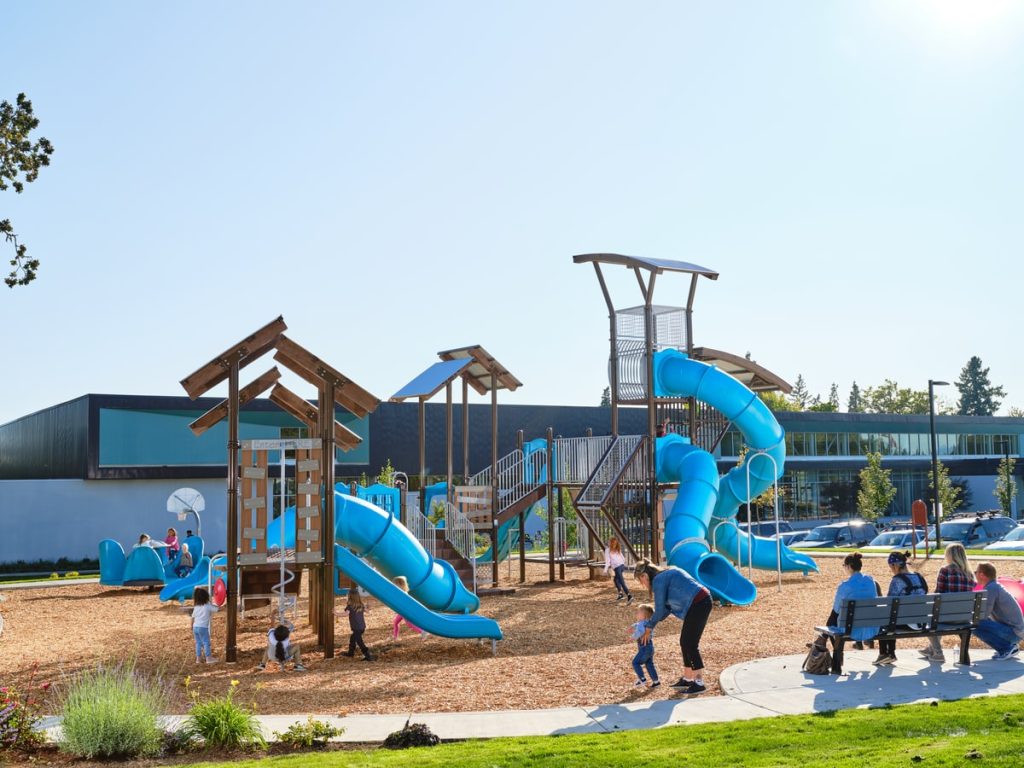 Blue tube slides on a wood-roofed play tower, with kids and parents on the wood chip surface.