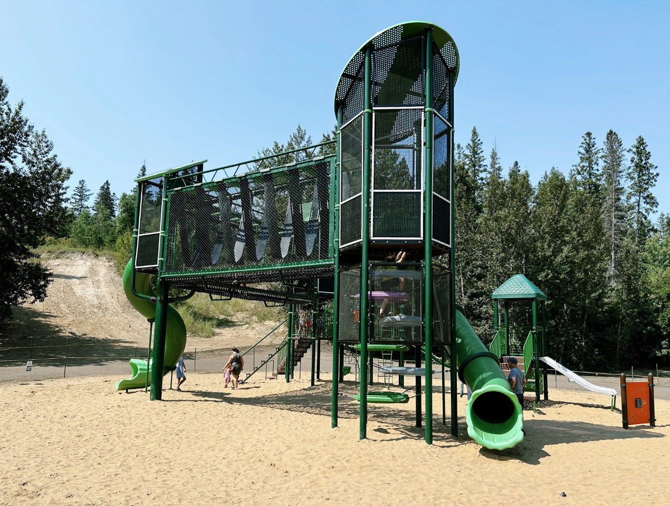 Green play tower with a tube slide and mesh-walled bridge on a sand surface, framed by a wooded hillside.