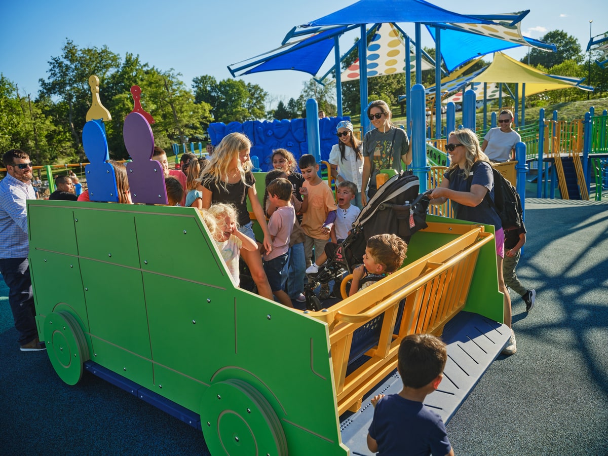 Children climbing into a green truck-shaped playground rocker beneath colorful shade sails.