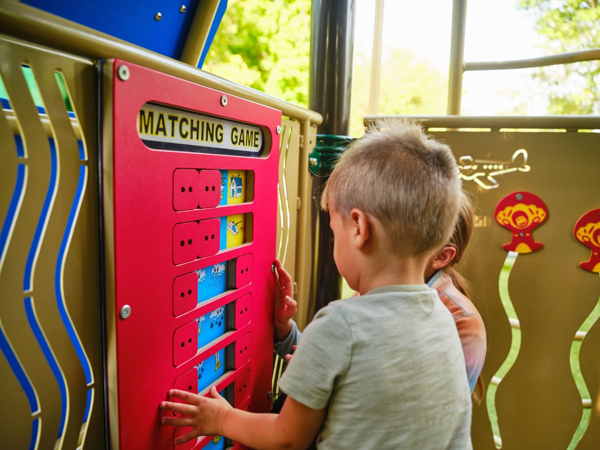 Child playing a matching game panel on a play structure, another child looking on.
