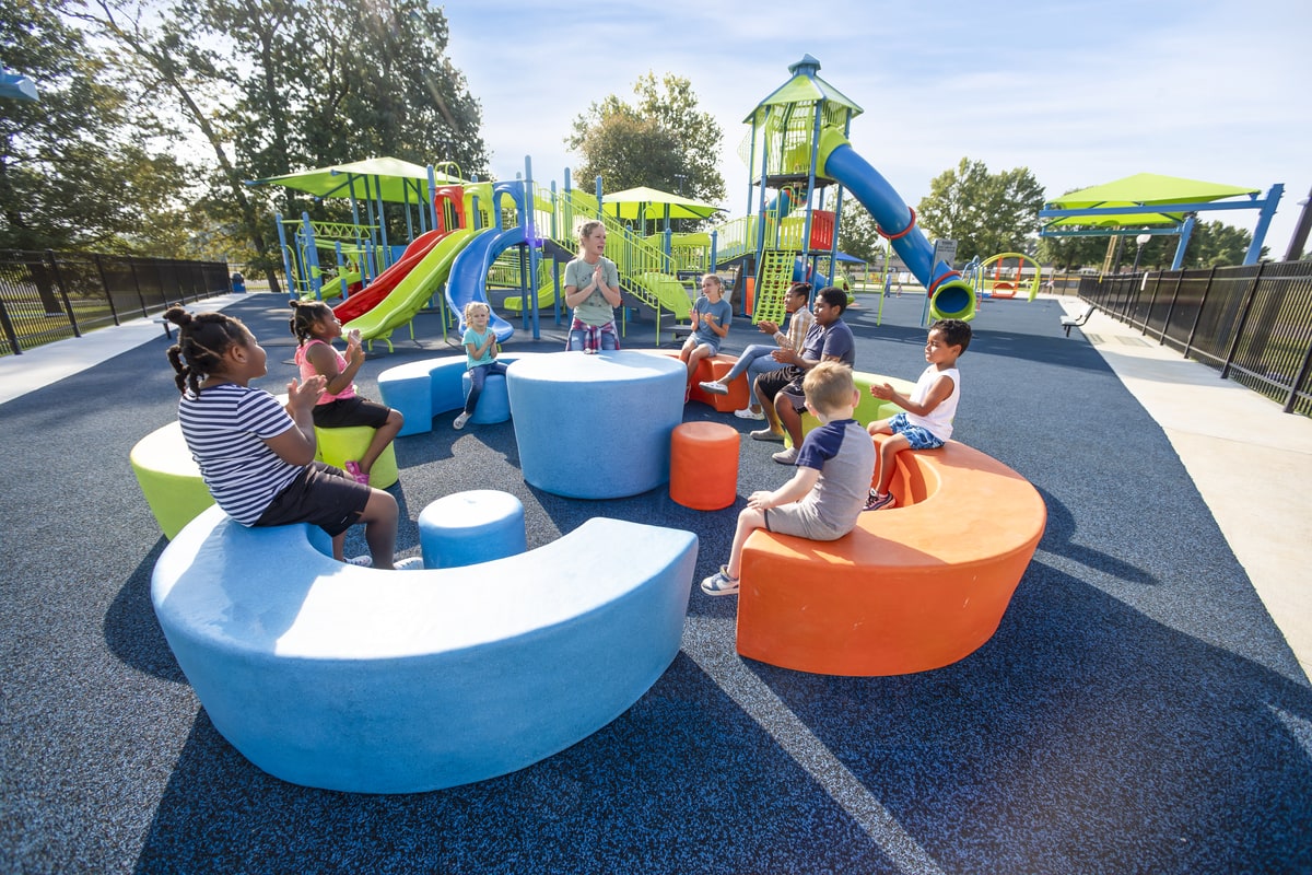 Children sitting on curved blue and orange benches near a green and blue play tower with a red slide.