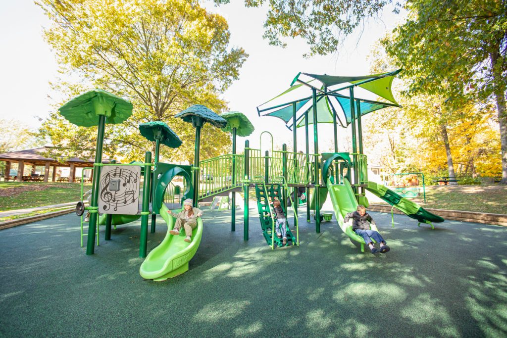 Green tower playground with a music panel and two slides, framed by autumn trees in a park.