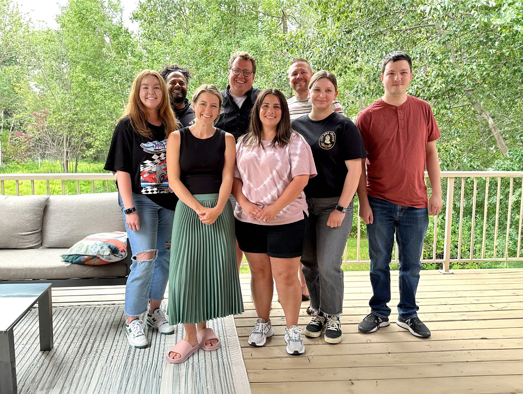 Eight team members smiling together on an outdoor deck surrounded by green trees.