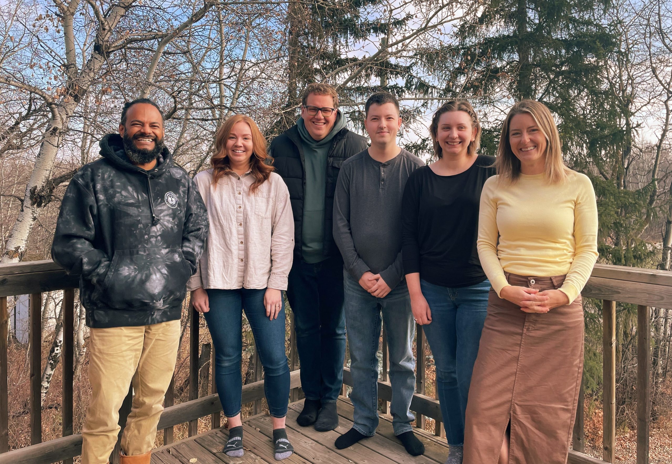 Group photo of six team members smiling together on an outdoor deck surrounded by trees.