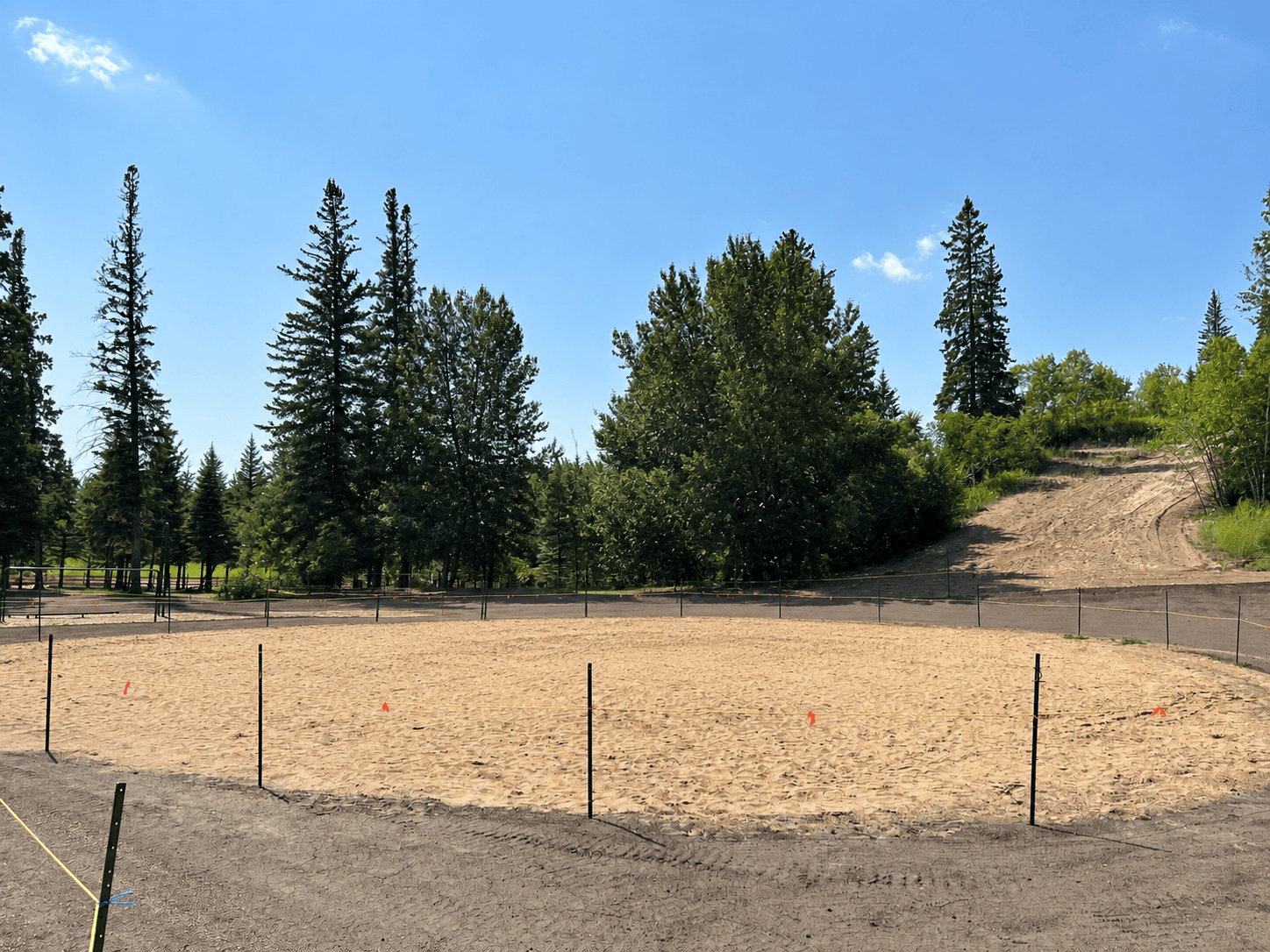 Empty sand play area marked with flags inside temporary fencing, framed by evergreen trees on a hillside.