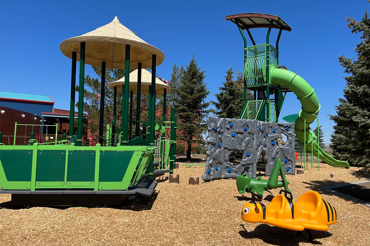 Green play tower with a spiral slide, rock climbing wall and a yellow bee spring rider on wood chips.