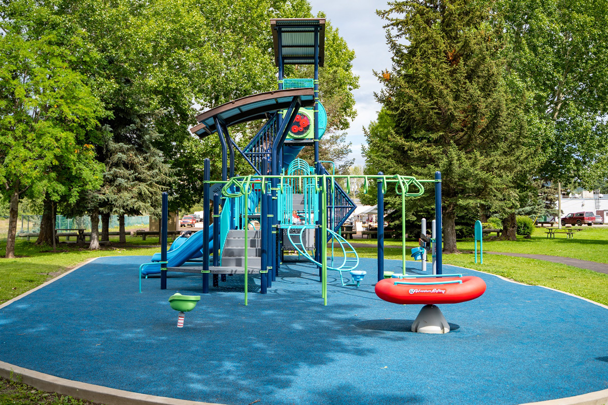 Teal play tower with a spiral slide, monkey bars and a red see-saw raft on blue rubber surfacing.