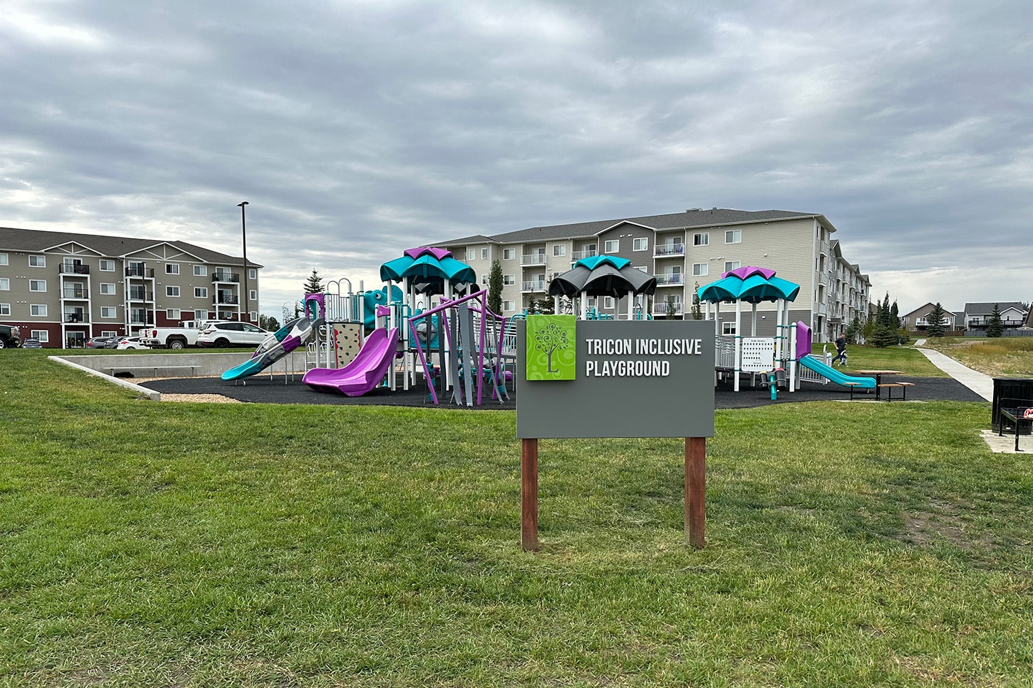 Tricon Inclusive Playground sign in front of a purple and teal accessible play structure near apartments.