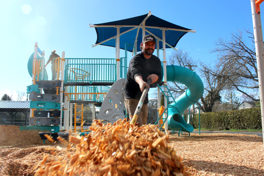 Worker shovels fresh wood chips around a teal play structure with a spiral slide and blue shade canopy.