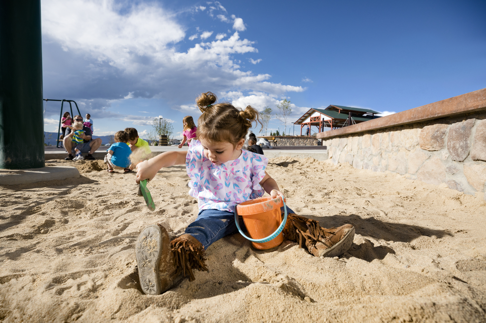 Toddler digs with a shovel and bucket in sand, one of the classic playground surfacing options, by a stone wall.