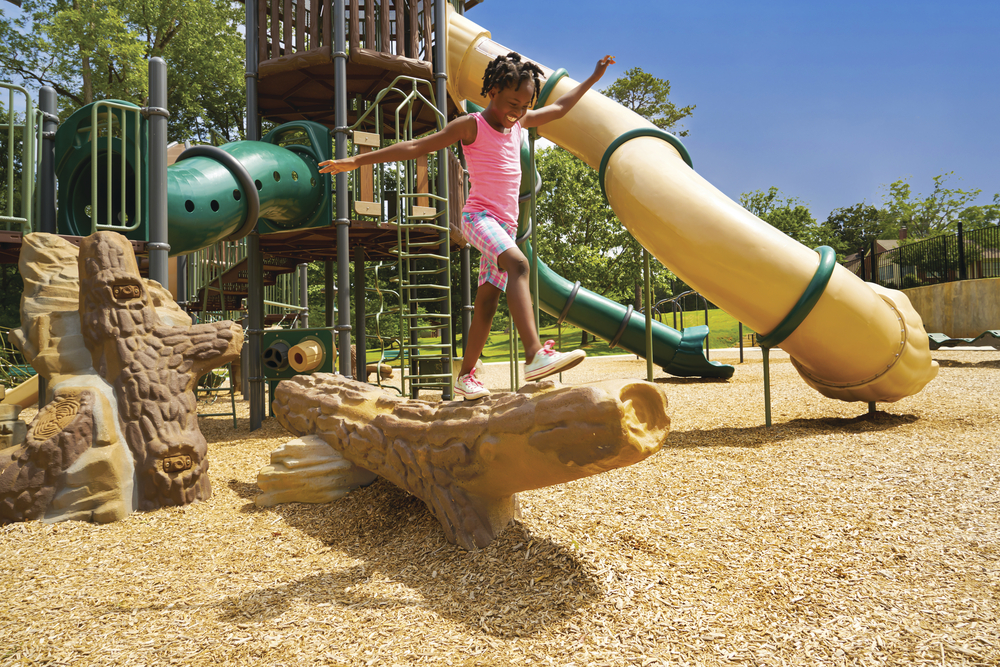 Girl balances on a log beam over wood chips, with a tan tube slide and green tunnel behind.