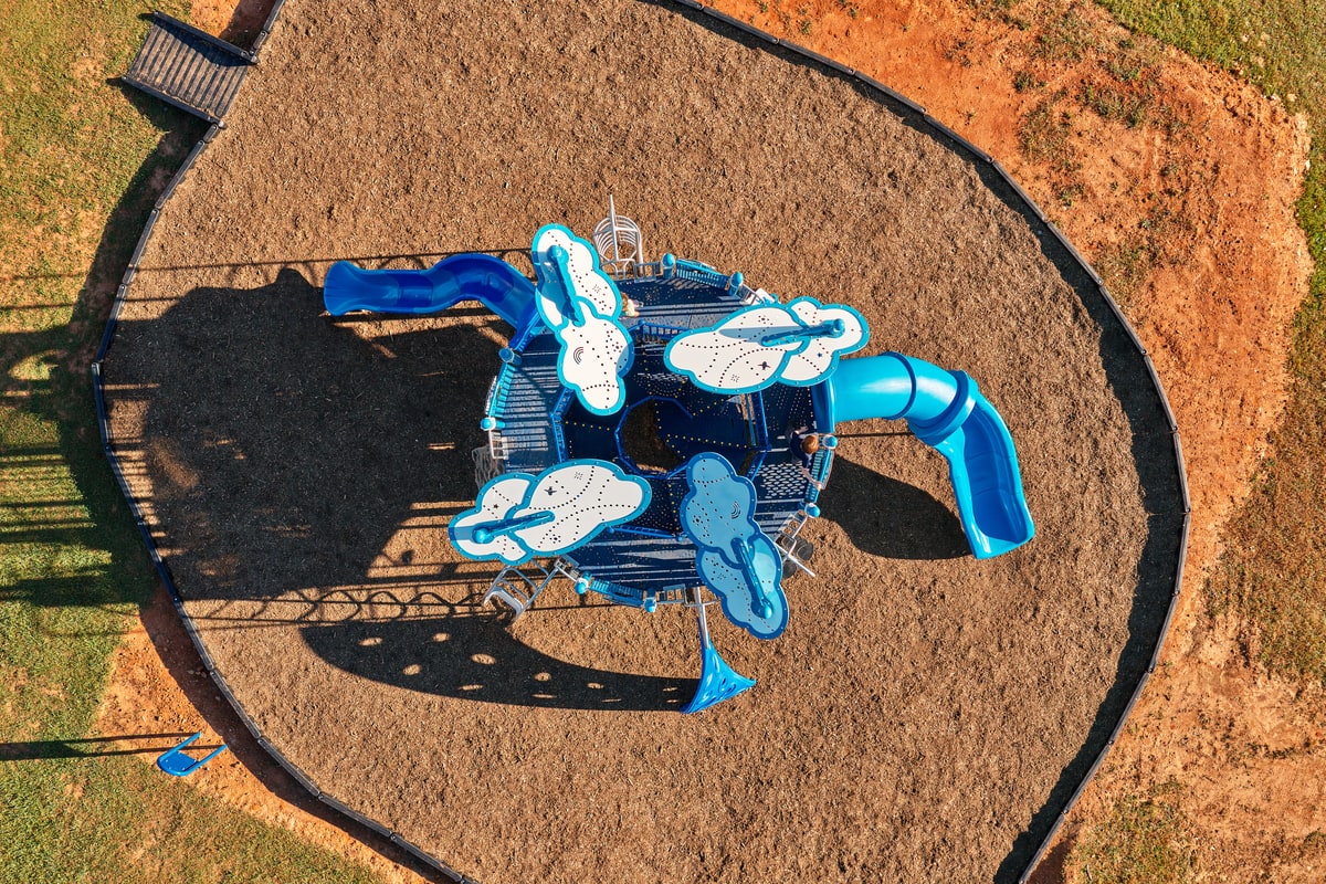 Aerial view of a blue play structure with cloud-shaped panels and two slides in a round mulch area.