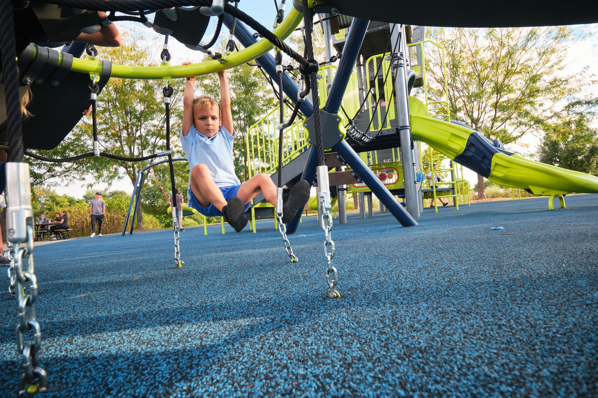 Boy hangs from a rope climber over blue poured rubber, with a lime slide and picnic tables behind.