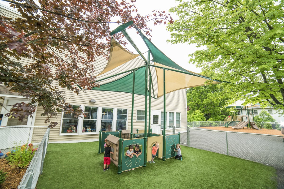 Toddlers explore playground equipment for small spaces, with play panels under shade sails in a fenced yard.