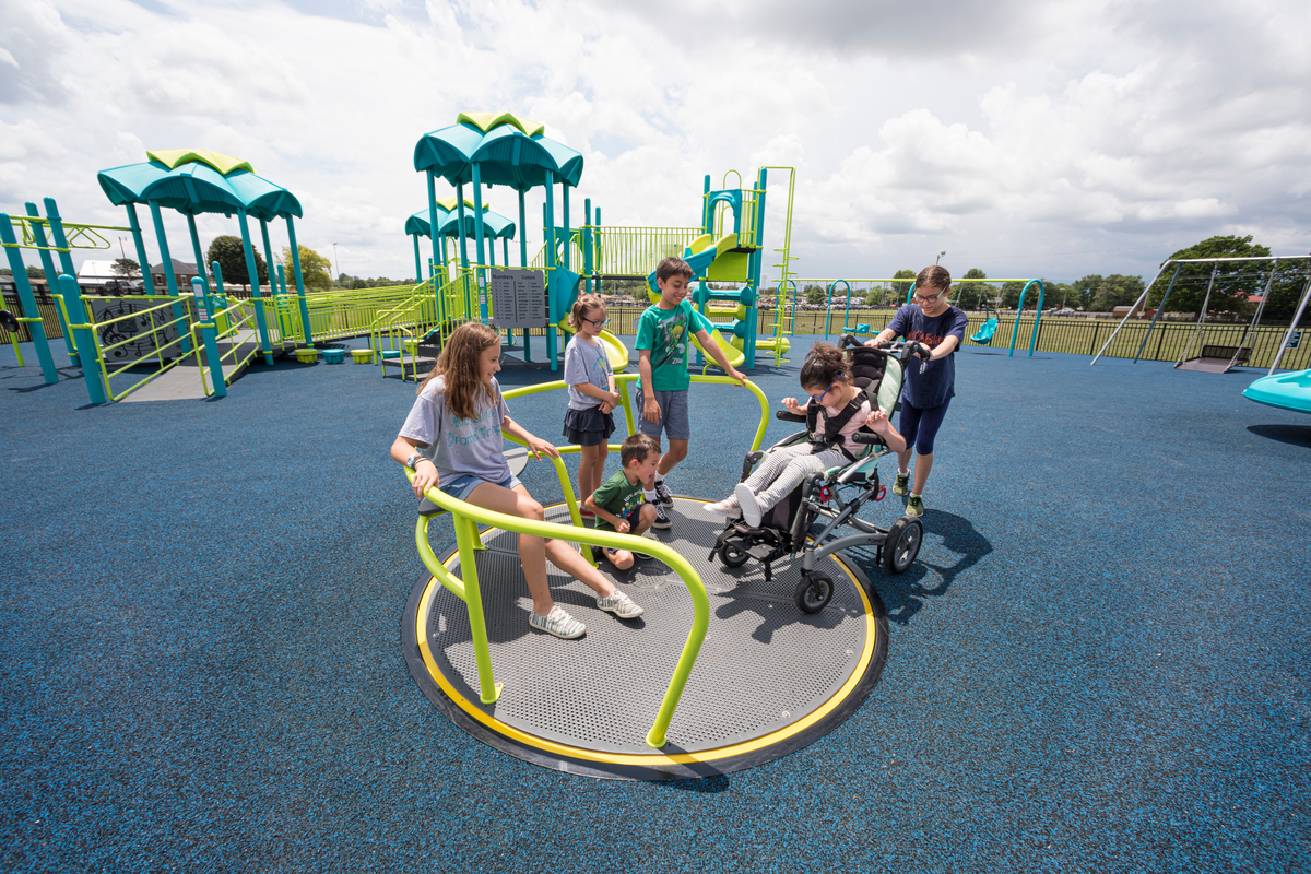 Children share an inclusive merry-go-round with a girl in a wheelchair on blue poured rubber surfacing.