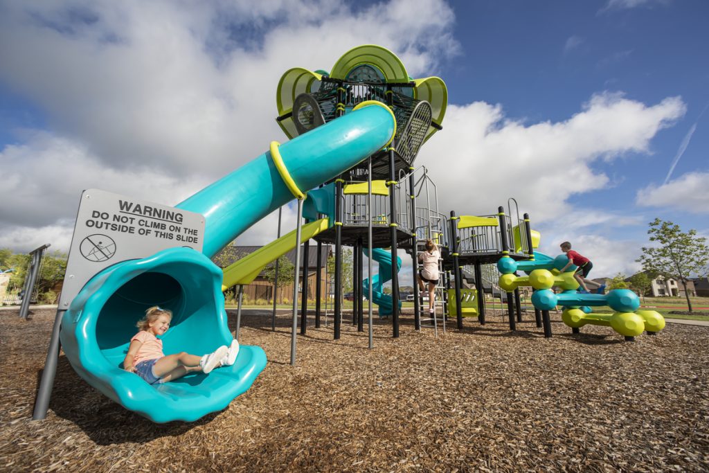Girl smiles at the bottom of a teal tube slide on wood chips, a warning sign and lime and teal tower behind.