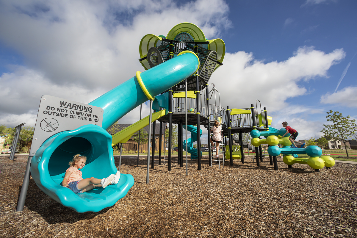 Girl smiles at the bottom of a teal tube slide on wood chips, a warning sign and lime and teal tower behind.