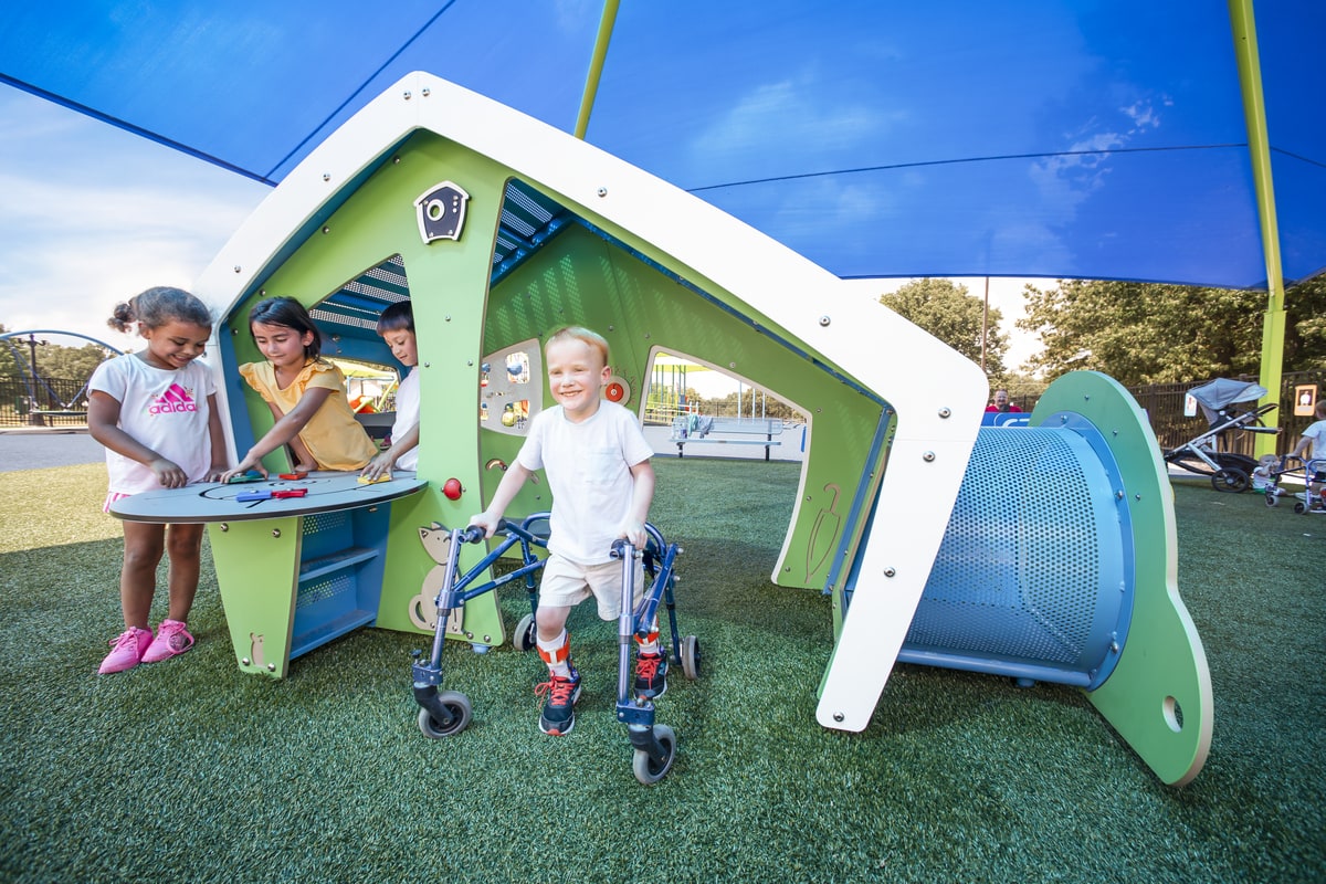 Boy using a walker beside an inclusive playhouse with a play table, children playing under a shade sail.