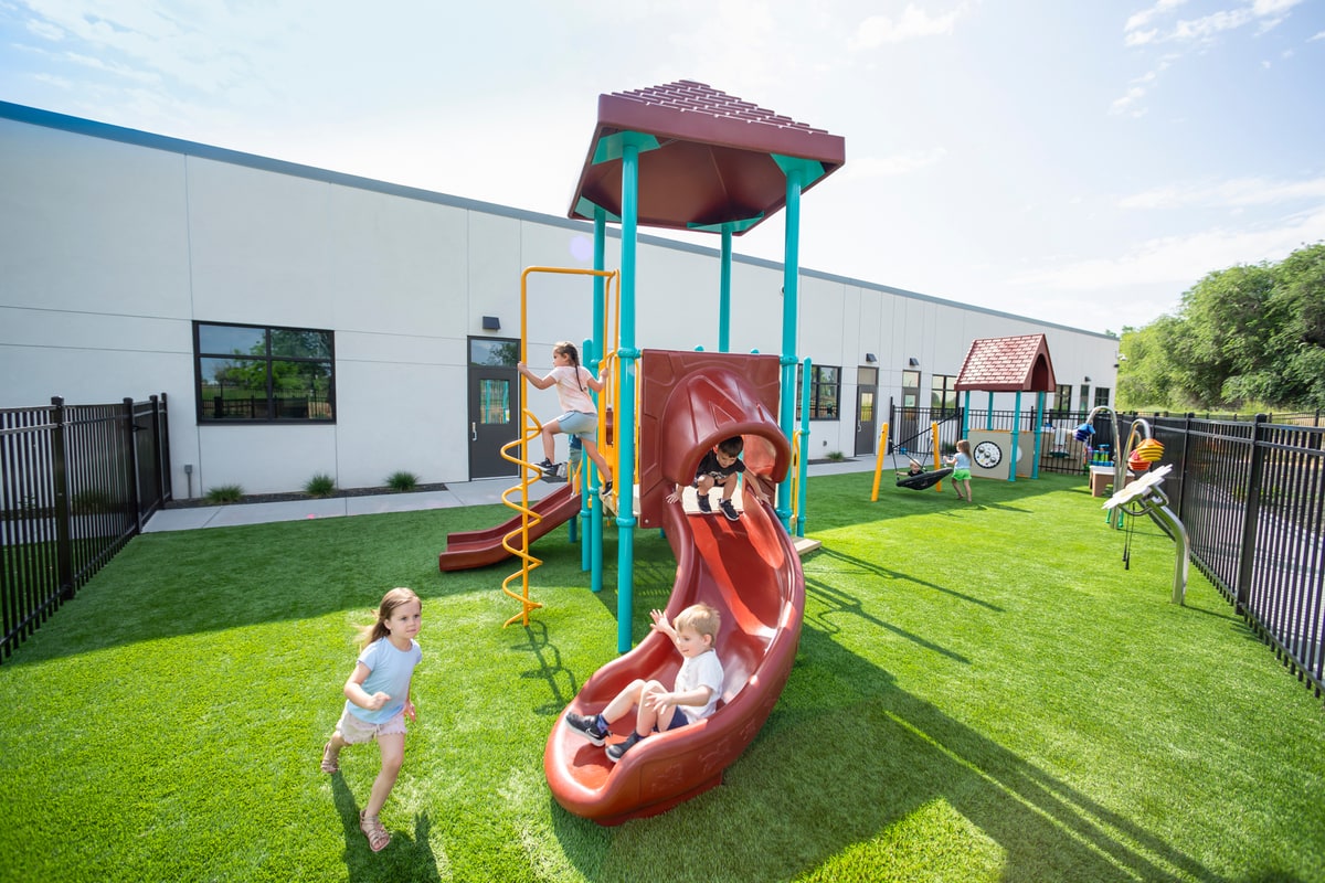 Children on a toddler structure with a curvy maroon slide and spiral climber on artificial turf.