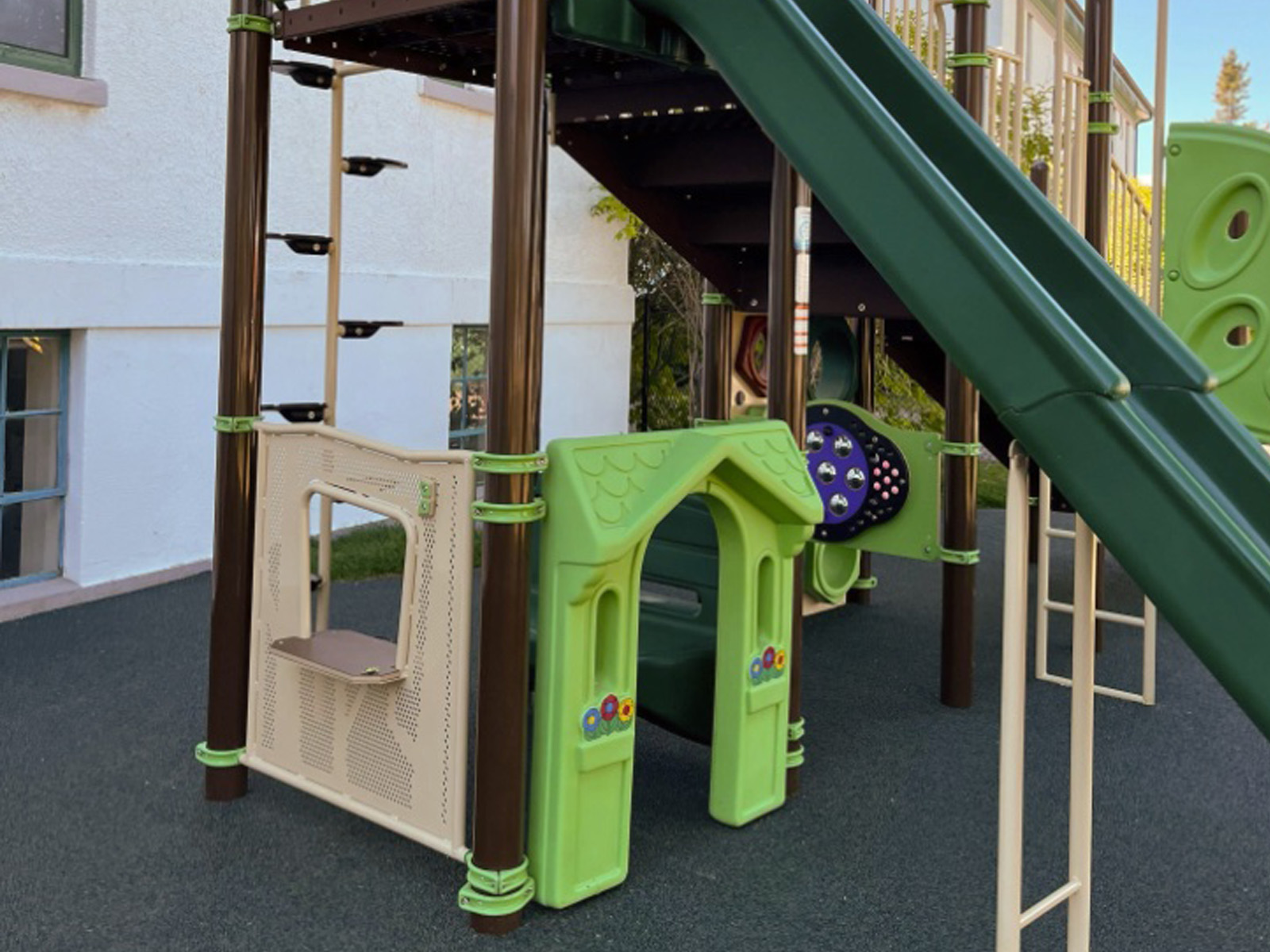 Lime playhouse arch, store counter panel and purple sensory panel beneath a green slide on rubber surfacing.
