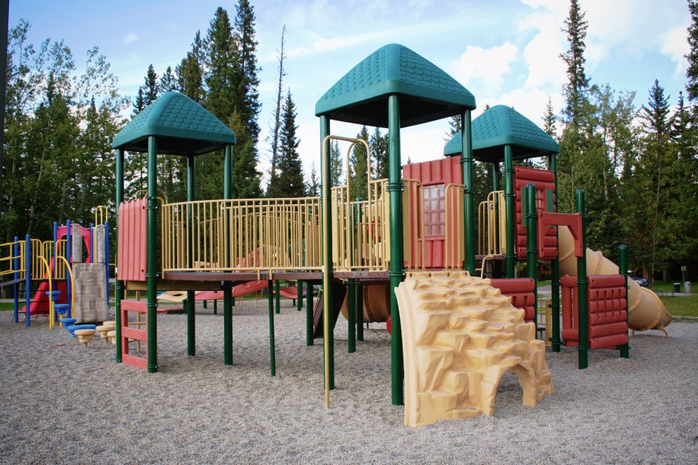 Green-roofed play structure with red and tan panels, a rock climber and tube slide on pea gravel, evergreens behind.