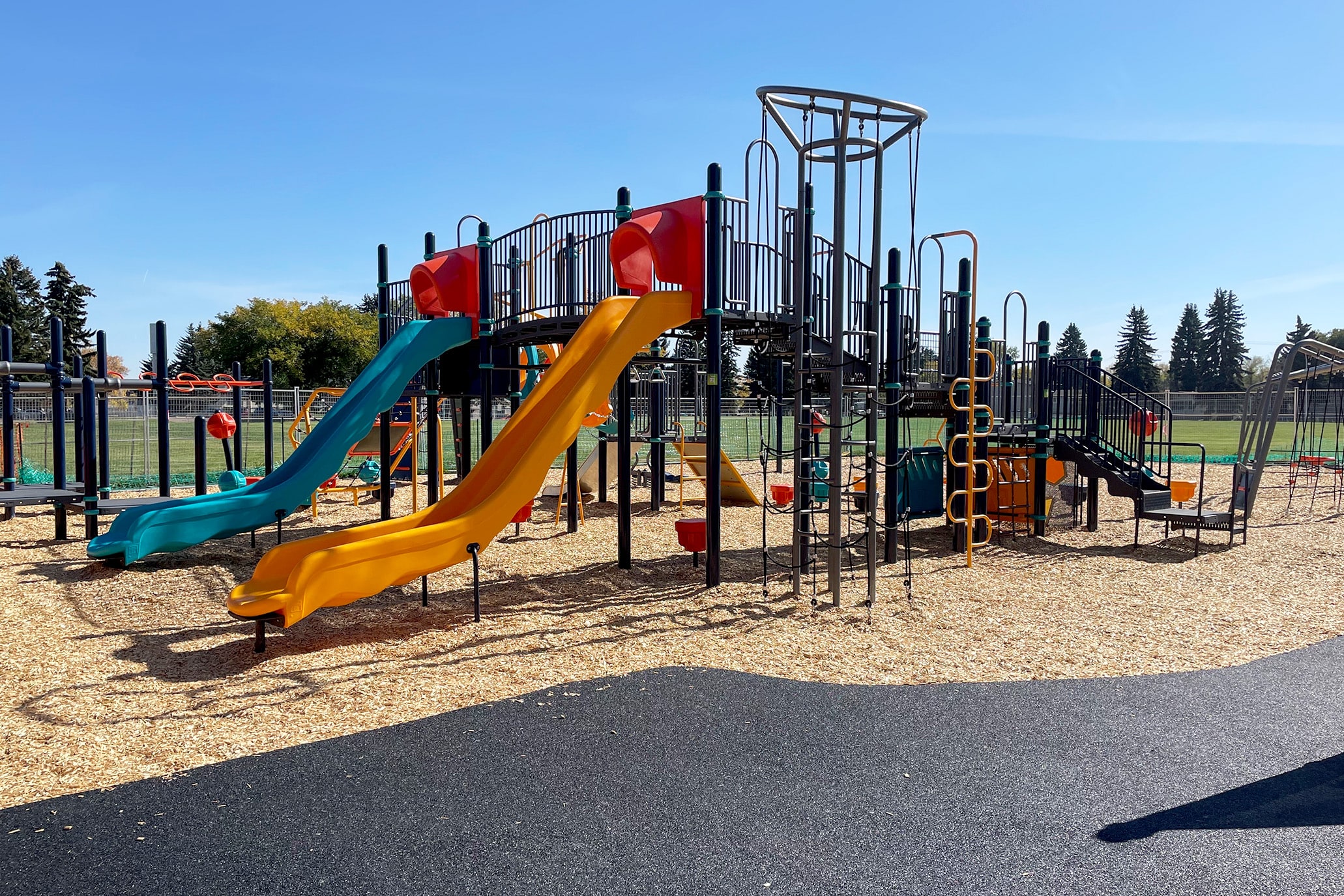Cobalt play structure with teal and yellow slides under orange hoods, set in wood chips beside a field.