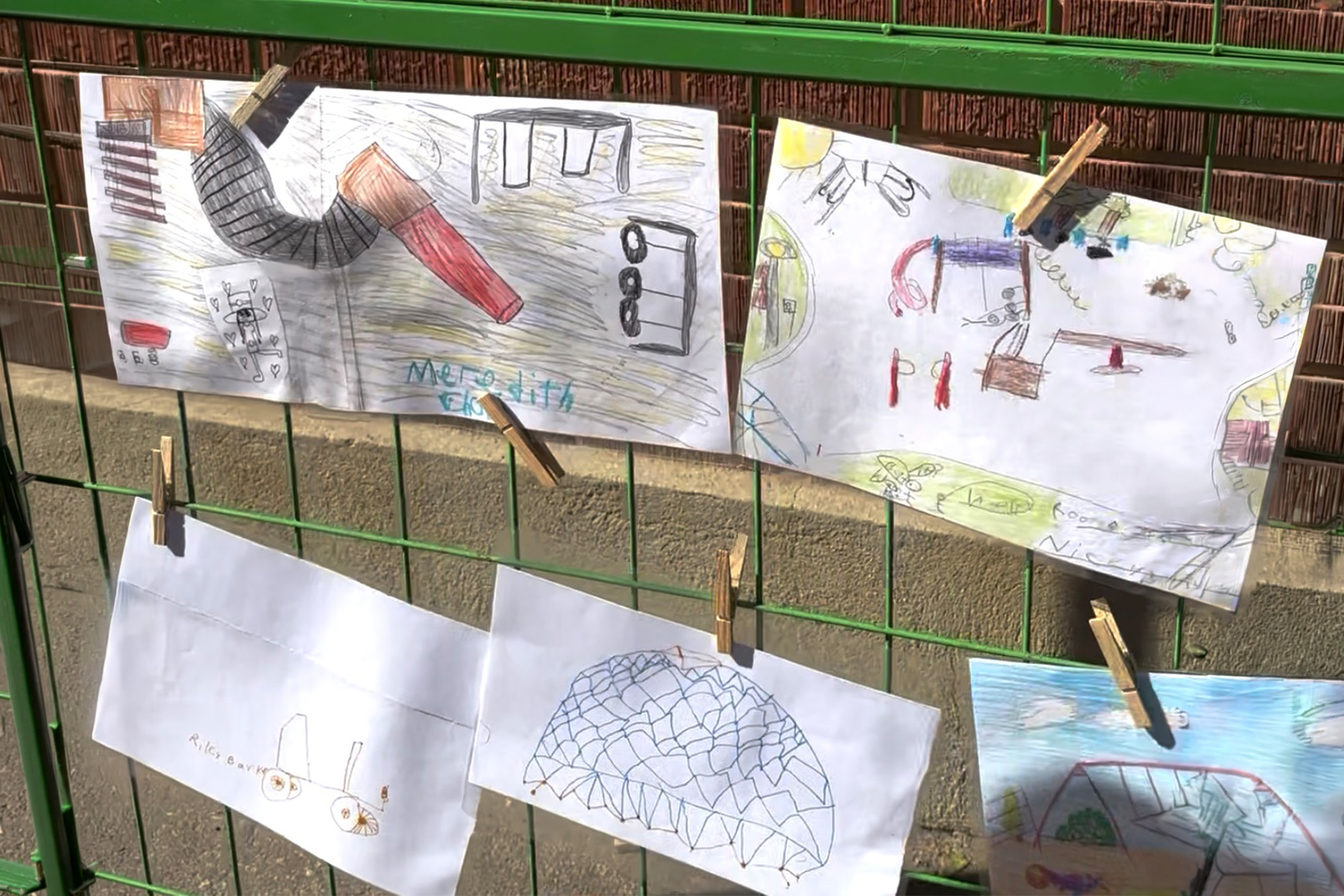 Students' drawings for school playground planning pinned to a fence, showing slides, nets and swings.