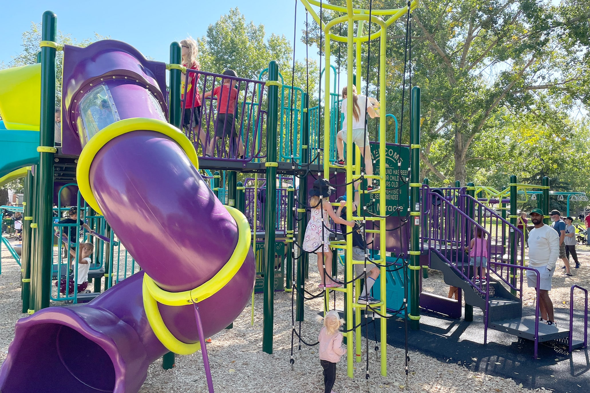 Children climb a lime rope tower beside a purple tube slide while an adult watches nearby.