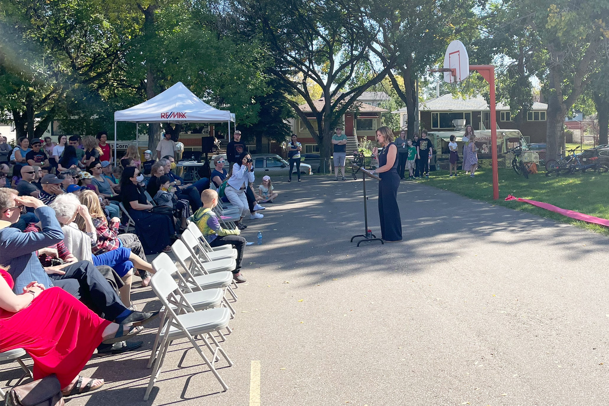 Speaker at a podium addresses a seated crowd at an outdoor playground opening celebration.